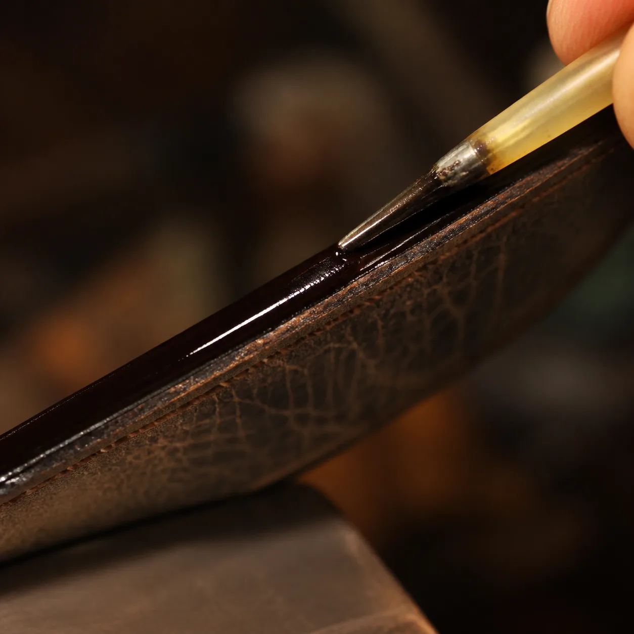 A fine applicator laying fresh glossy paint along the worn edge of a leather strap